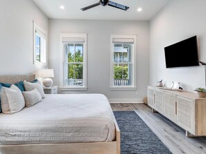 A bedroom with a large bed, two windows, a ceiling fan, a TV, and a wooden cabinet. - Honeybee | Elegant 30A Retreat, 6 Bikes +Community Pool (Santa Rosa Beach)