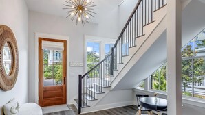 Un intérieur moderne avec un escalier, une porte en bois, une table ronde entourée de chaises et une vue sur l’extérieur.