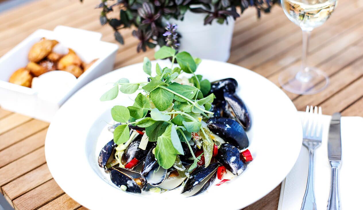 Un plato de mejillones con verduras y pimientos rojos, una copa de vino blanco y un bol pequeño de alimentos fritos sobre una mesa de madera.
