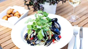 Un plato de mejillones con verduras y pimientos rojos, una copa de vino blanco y un bol pequeño de alimentos fritos sobre una mesa de madera.