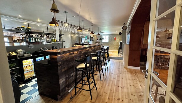 Zona de bar con suelo de madera, taburetes, una barra con encimera de madera y luces colgantes.