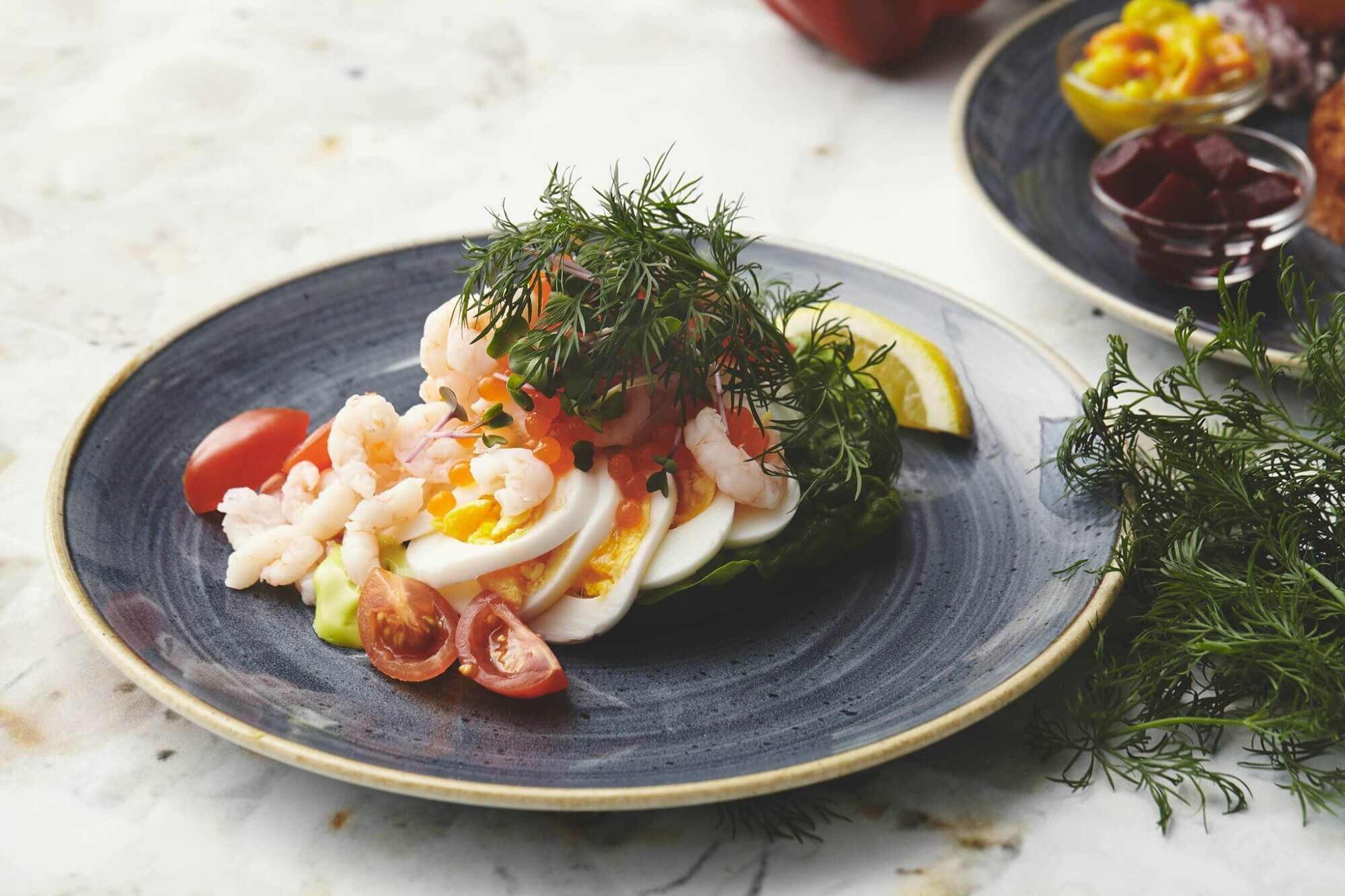 Un plato de comida con camarones, huevos y una rebanada de aguacate, adornado con eneldo y limón.