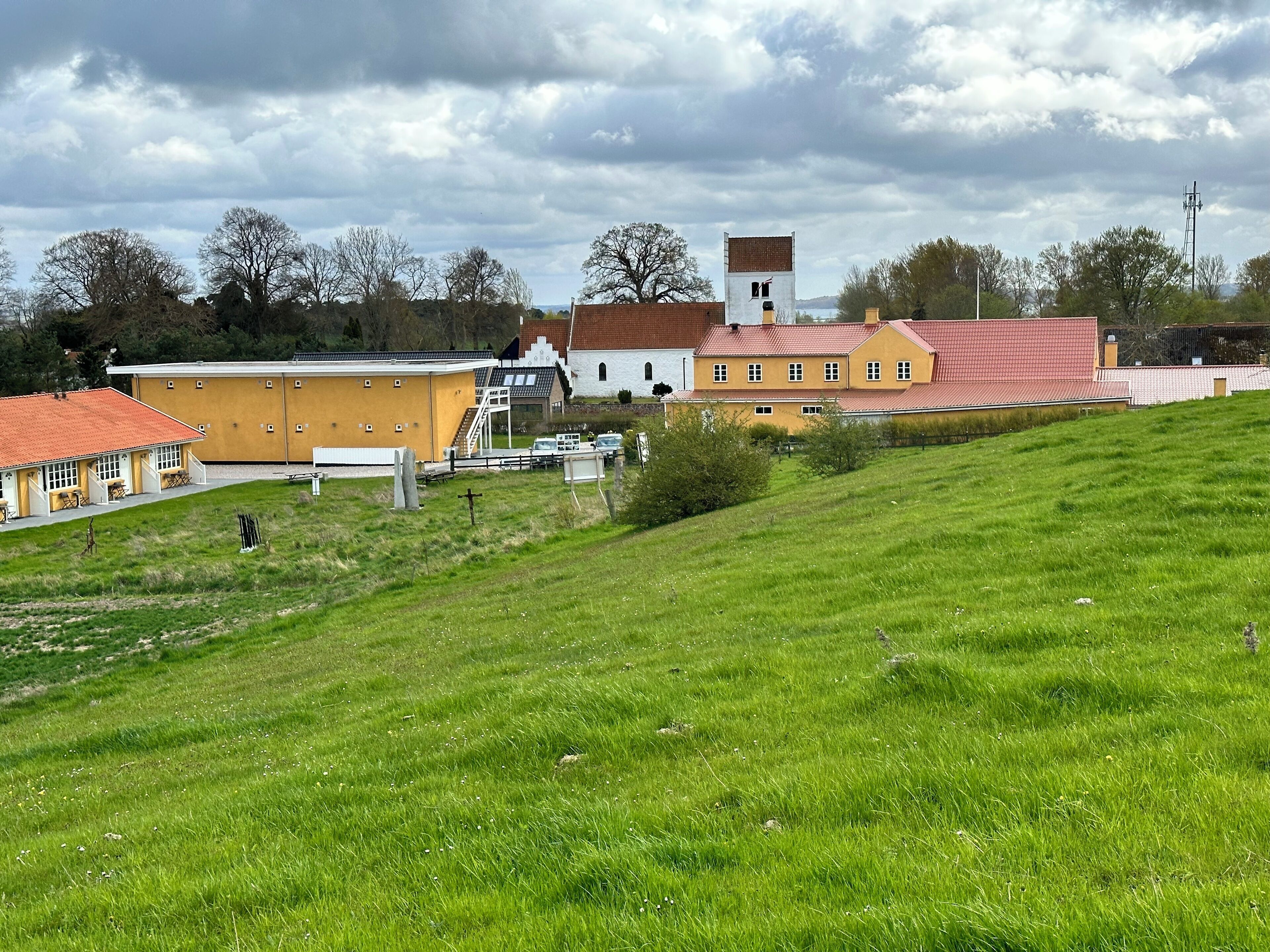 A rural setting with a yellow building, a white church with a red roof, and a hill with green grass.