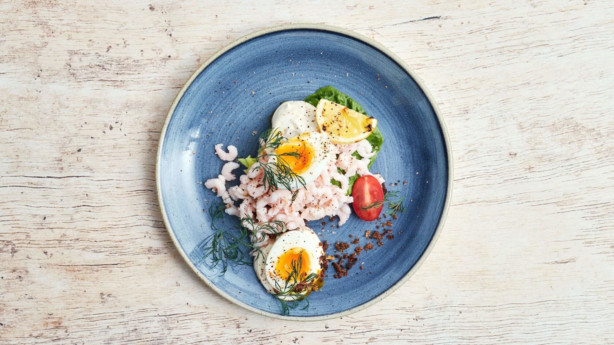 Une assiette bleue contenant des crevettes, des œufs et des légumes.