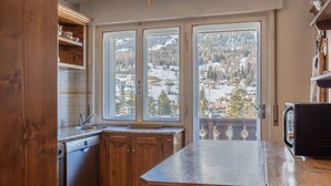 A kitchen with wooden cabinets, a dishwasher, and a large window offering a view of a snowy landscape. - Casa Cristallino, in the town center with panoramic view (Cortina d´Ampezzo)