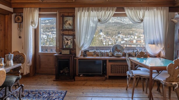 A cozy room with wooden walls and ceiling, a large window with curtains, a wooden table with chairs, a fireplace, and a view of a snowy village. - Casa Cristallino, in the town center with panoramic view (Cortina d´Ampezzo)