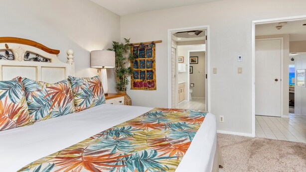 Quarto de hotel com cama, mesa de cabeceira, abajur, tapeçaria na parede e vista para o mar por uma porta aberta.