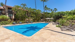 Piscina com espreguiçadeiras, cercada por palmeiras e um edifício com varandas.