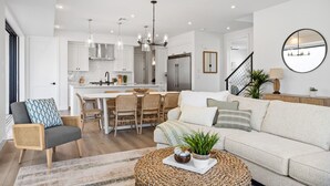 A modern living room with a sofa, a coffee table, and a dining area with chairs and a kitchen in the background. - Sunstone 6 Bedroom Home (St. George)