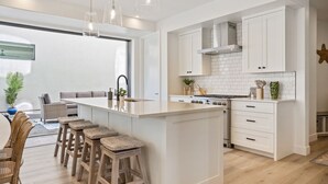 A modern kitchen with white cabinetry, a central island with stools, a built-in stove, and pendant lighting. - Sunstone 6 Bedroom Home (St. George)