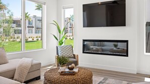 A modern living room with a large window, a flat-screen TV mounted on the wall, and a fireplace below it. - Sunstone 6 Bedroom Home (St. George)