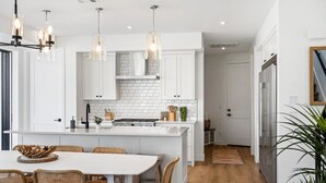 A modern kitchen with white cabinetry, a central island, and a dining area with wicker chairs. - Sunstone 6 Bedroom Home (St. George)
