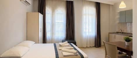 Una habitación de hotel con cama, escritorio, silla, kitchenette y cortinas.
