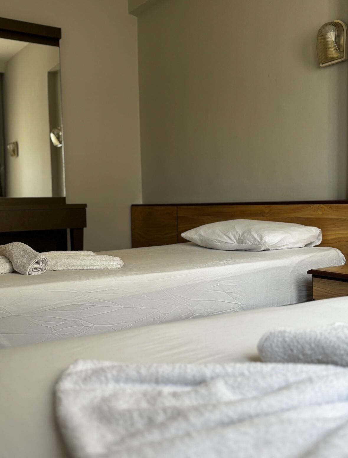 A hotel room with two beds, a wooden headboard, a nightstand, a mirror, and a wall-mounted light fixture.