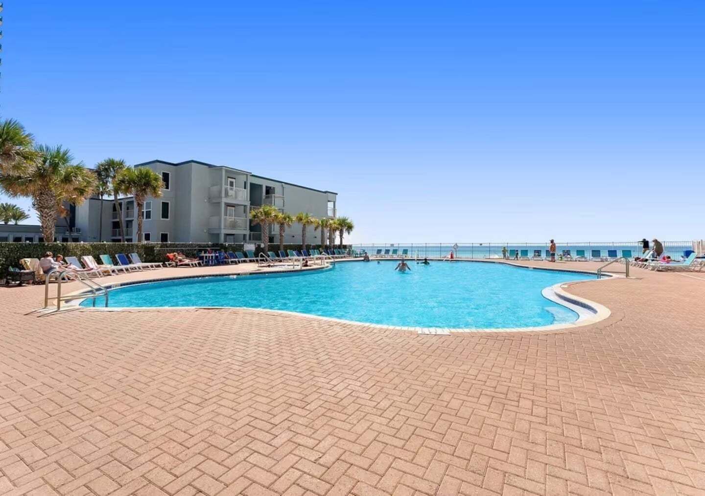 A swimming pool with lounge chairs, a building with balconies, and a clear sky.