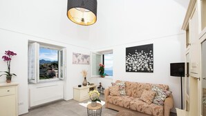 A modern living room with a sofa, a round ottoman, a small side table with a vase, and a window with a view of the outdoors. - SAM'S HOUSE - A corner of paradise suspended between sky and sea (Amalfi)