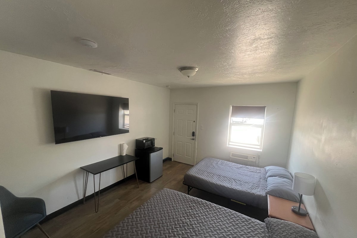 A compact room with a flat-screen TV, a desk, a chair, two beds, a window, and a ceiling light.