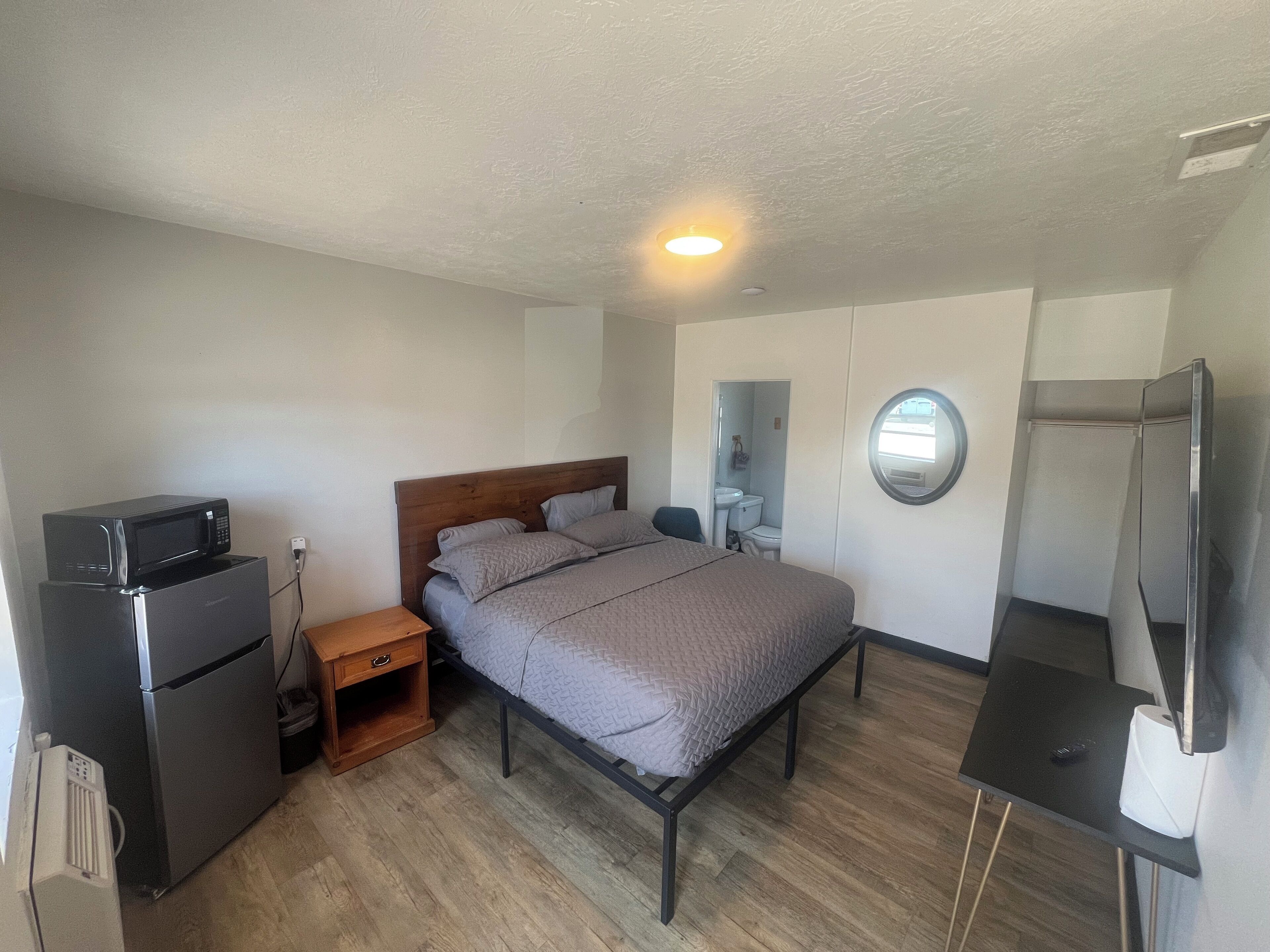 A compact bedroom with a bed, a small wooden nightstand, a built-in microwave, a toilet, a sink, and a television.