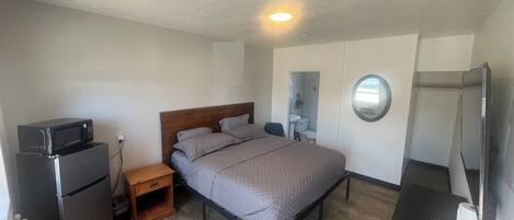 A compact bedroom with a bed, a small wooden nightstand, a built-in microwave, a toilet, a sink, and a television.