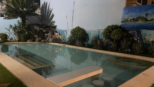 Une piscine bordée de pierres, un espace détente avec des tables rondes, et une fresque représentant un paysage tropical.