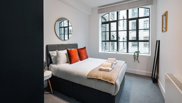 Une chambre moderne avec un grand lit, un miroir rond, une fenêtre donnant sur des immeubles et une table de chevet.