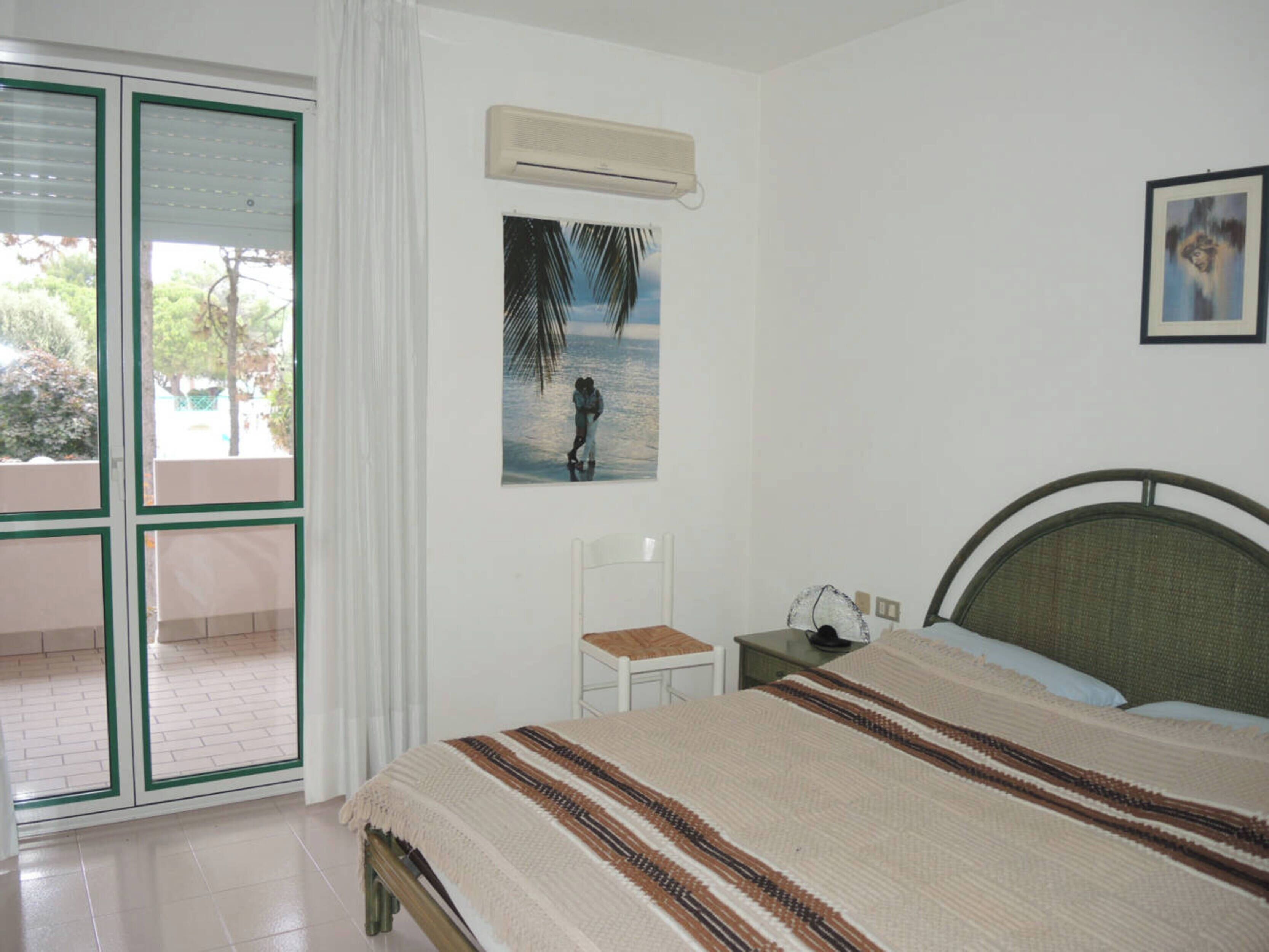 A bedroom with a bed, a nightstand, a chair, a picture on the wall, and an air conditioning unit.