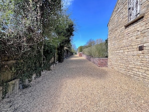 Un chemin de gravier mène à un bâtiment en pierre, doté d’un mur en briques et d’une clôture en bois.