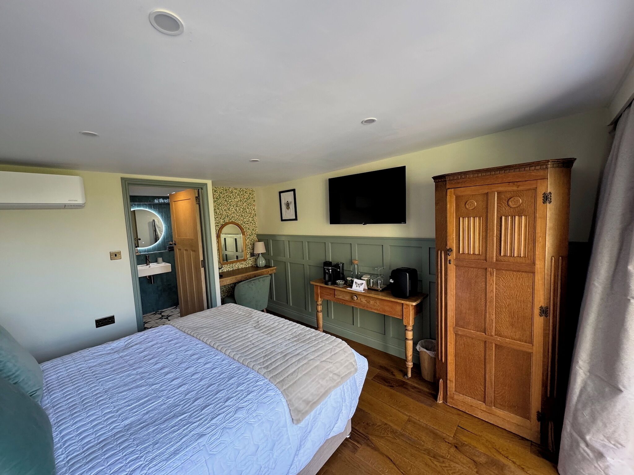 Un dormitorio con cama, un armario de madera, un escritorio con espejo, televisión y un baño visible a través de una puerta abierta.