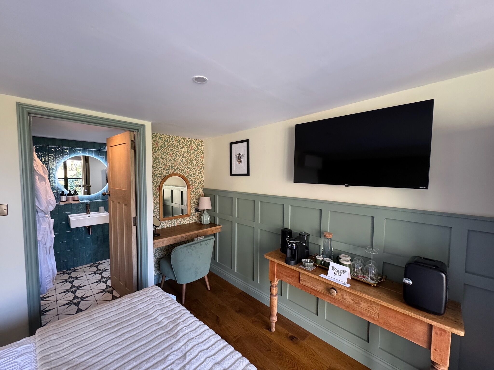 A hotel room with a bed, a desk with a computer, a TV, a bathroom with a mirror, and a shower.