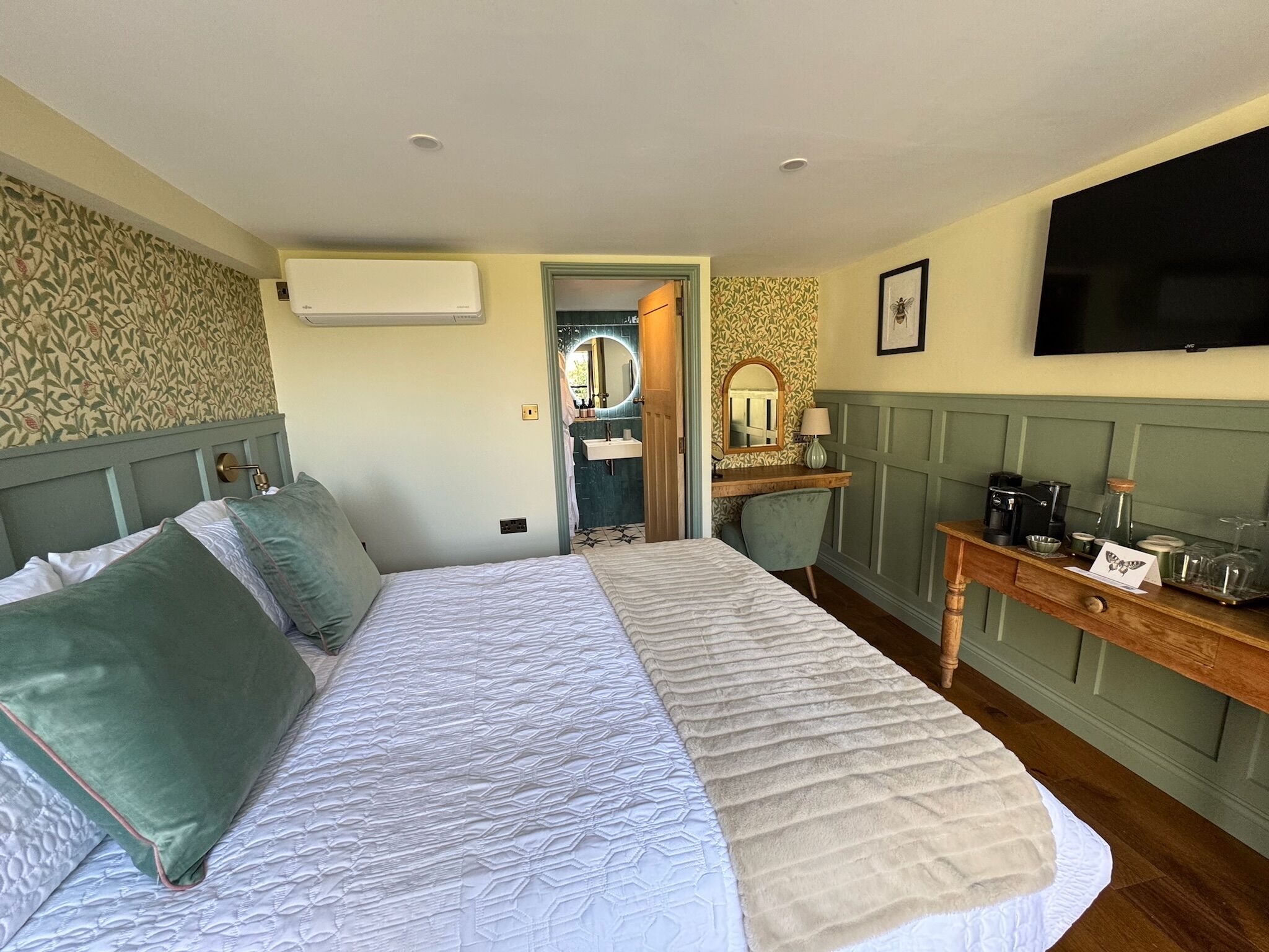 A bedroom with a large bed, a bedside table with a coffee maker, a mirror, and a TV on the wall.