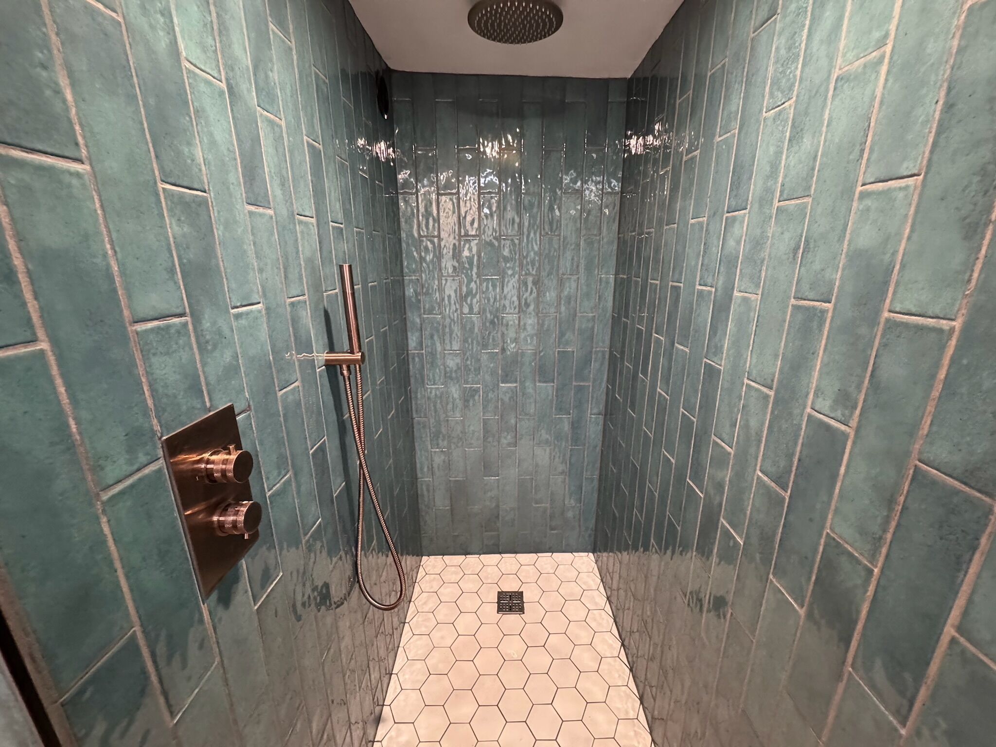 Eine Dusche mit Handbrause und Wandarmatur, umgeben von wandbündig in Türkis gefliesten Wänden und einem Boden mit Sechseckfliesen.
