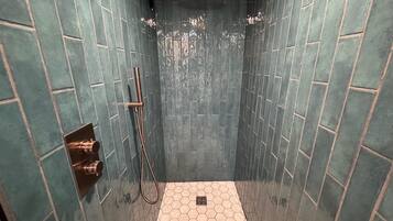 Eine Dusche mit Handbrause und Wandarmatur, umgeben von wandbündig in Türkis gefliesten Wänden und einem Boden mit Sechseckfliesen.