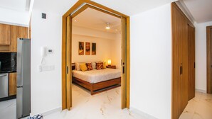 Een moderne slaapkamer met een houten bed, een nachtkastje en een slaapkamertje. Er is een keukenruimte met een koelkast en kasten zichtbaar door een open doorgang.