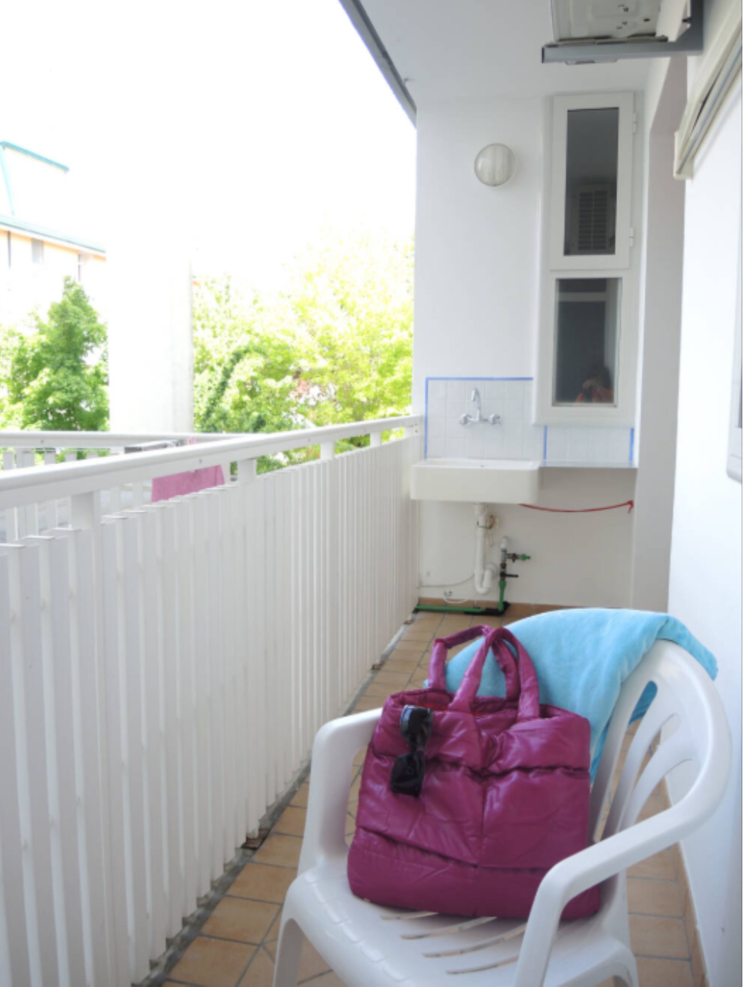 Un balcón con una silla blanca y un bolso rosa, un lavamanos blanco y una ventana con vistas a la vegetación.
