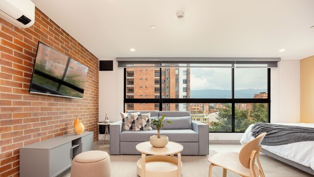 A modern hotel room with a brick accent wall, a grey sofa, a round wooden table, a wooden chair, and a large window offering a city view.