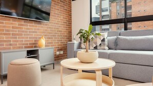 A modern living room with a grey sofa, a round wooden coffee table, a beige ottoman, a large flat-screen TV mounted on a brick wall, and a potted plant on the coffee table.