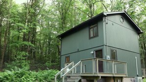Eine zweistöckige Holzhütte mit Balkon und Treppe, umgeben von dichtem Wald.