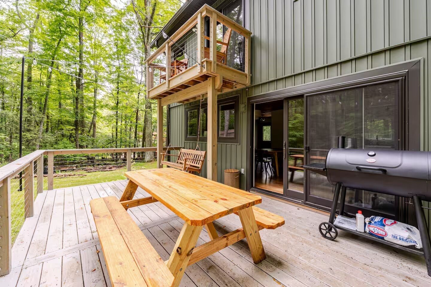 Une terrasse en bois avec une table de pique-nique et un barbecue, entourée d’une forêt.