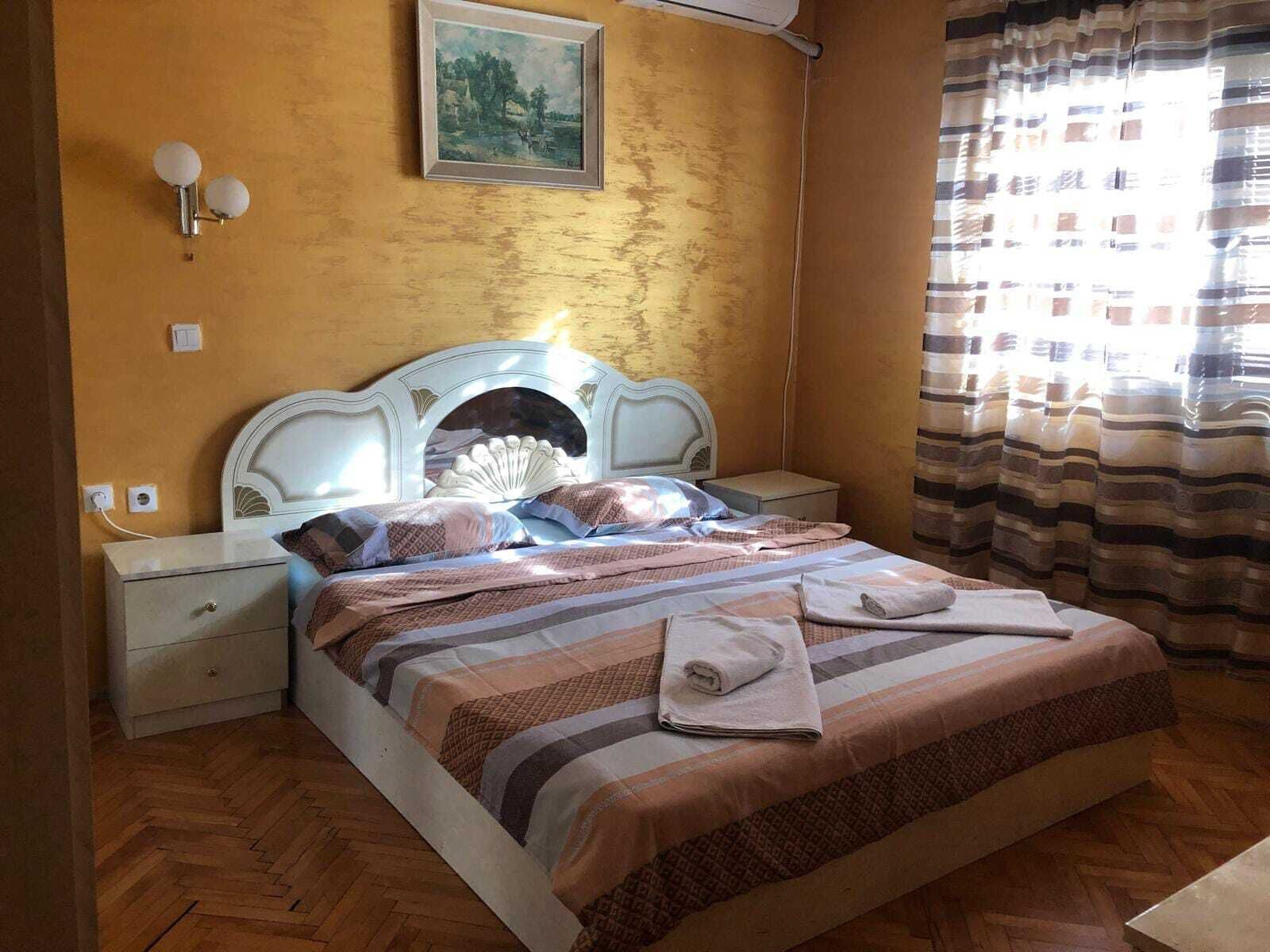 A bed with a patterned bedspread, a nightstand with a lamp, a wall-mounted picture, and a window with curtains.