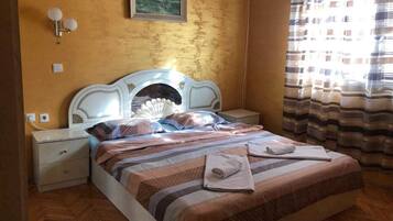 A bed with a patterned bedspread, a nightstand with a lamp, a wall-mounted picture, and a window with curtains.