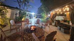 An outdoor patio with a fire pit surrounded by wooden chairs, a covered seating area with string lights, and tropical plants.