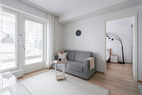 A modern living room with a grey sofa, a small round table with a book and a vase, a clock on the wall, and a large window with curtains. - Modern 1BR Apartment in Kaleva (Tampere)