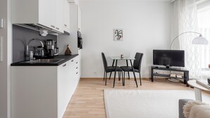 Eine moderne Küche mit weißen Schränken, einer schwarzen Arbeitsplatte und einem Essbereich mit einem Glastisch und schwarzen Stühlen. Ein Fernseher steht auf einem Schrank und ein Fenster ist mit transparenten Vorhängen drapiert.