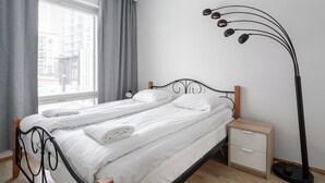 Ein ordentlich bezogenes Doppelbett mit weißen Laken, ein Nachttisch mit Schublade und eine Stehlampe.