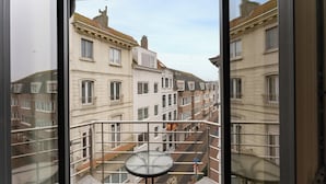 Un balcon avec une table et des chaises en verre, donnant sur une rue bordée d’immeubles.
