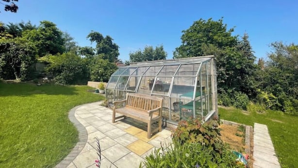 A greenhouse with a bench and a table, surrounded by a garden with various plants and a clear blue sky. - Bungalow with distant sea views (Colwyn Bay)