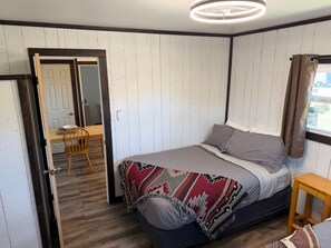 A bedroom with a bed, a wooden chair, a small wooden table, and a door leading to another room.