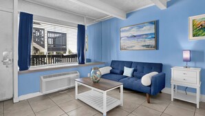 A room with a blue sofa, a white side table, a painting of a beach scene, and a window with blue curtains.