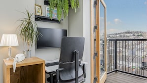 A modern office space with a desk, chair, and computer. There is a potted plant, a lamp, and a mounted TV. The room has a large window with a view of a cityscape. - Luxury Loft Living | Free Parking - Pass the Keys (Brighton)