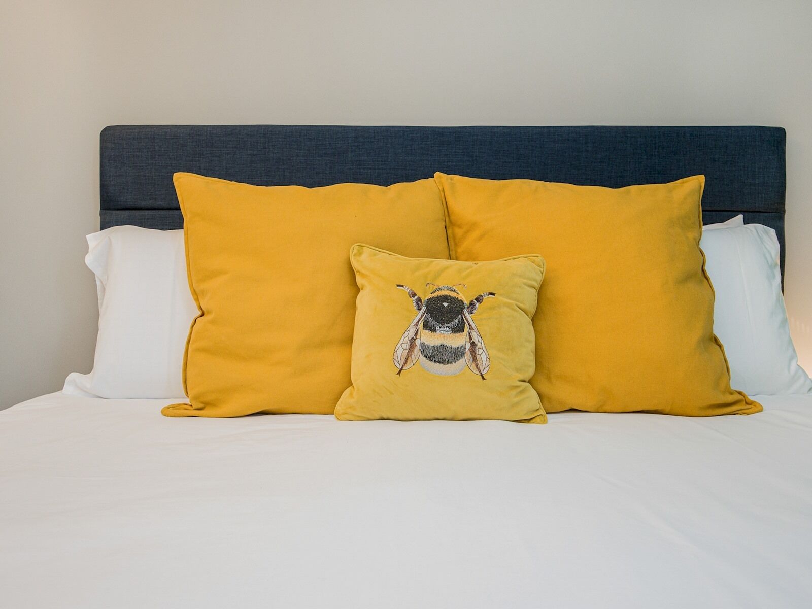 Un lit avec une tête de lit sombre, deux grands oreillers jaunes et un plus petit oreiller orné d’un motif d’abeille.